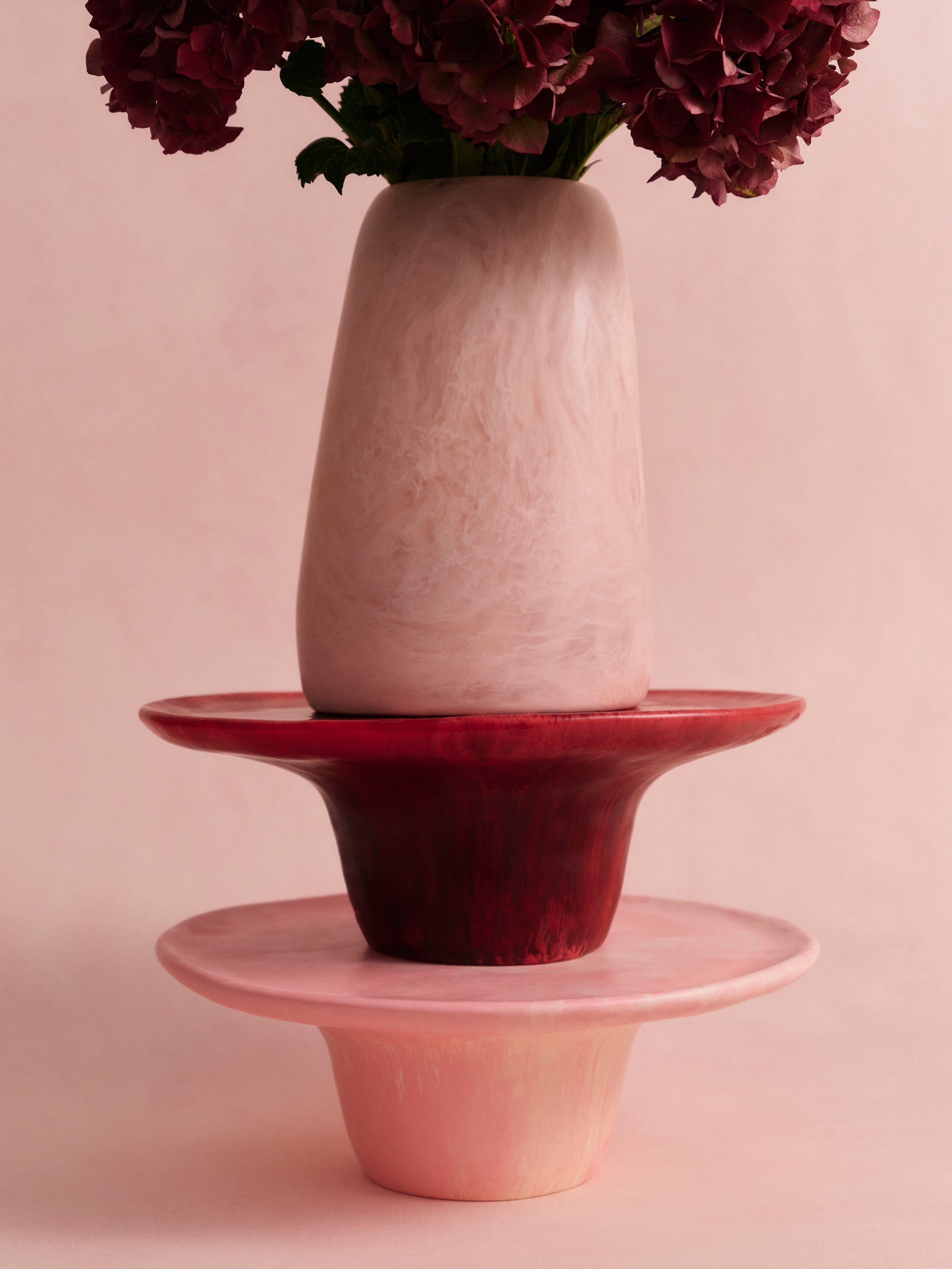 Two pink cake stands with a large pink resin vase atop with deep pink flowers on a light pink backdrop